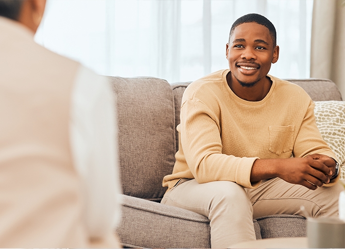 A smiling man sitting on a couch talking with another person.